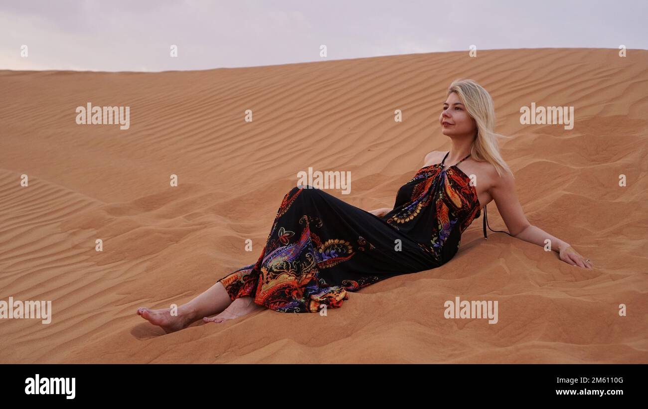 A young blonde lies on a pesce in the desert. Woman in the dunes. Travel and beauty. Stock Photo