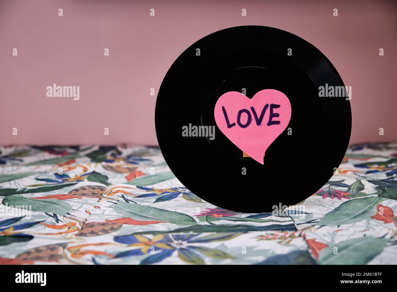 Black vinyl record with a heart in the centre above a table with a tropical pattern and a pink background. Stock Photo