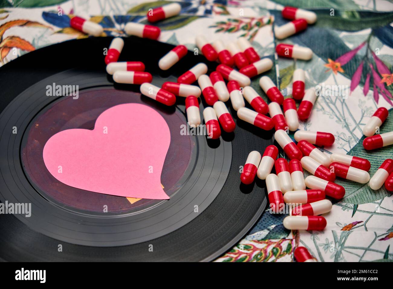 Vinyl record with a pink heart and a bunch of pills, on top of a table with a tropical pattern. Stock Photo