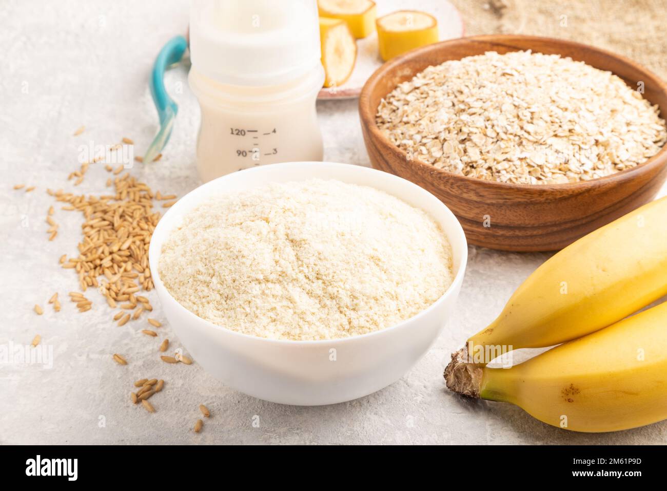 Powdered milk and oatmeal, banana baby food mix, infant formula, pacifier, bottle, spoon on gray concrete background. Side view, close up, artificial Stock Photo