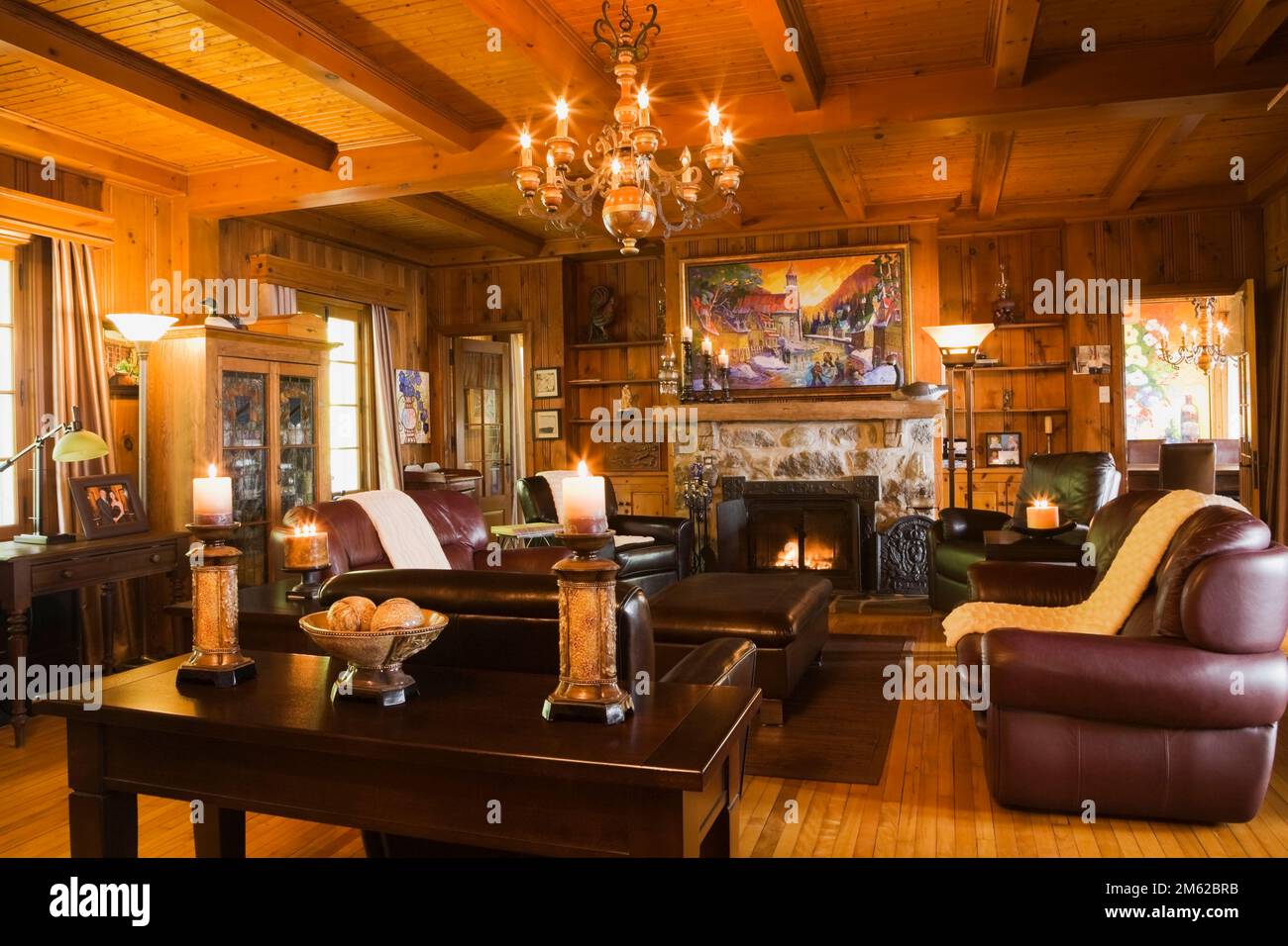 Lit natural stone fireplace and brown leather armchairs and sofas in living room inside old 1920s cottage style home. Stock Photo