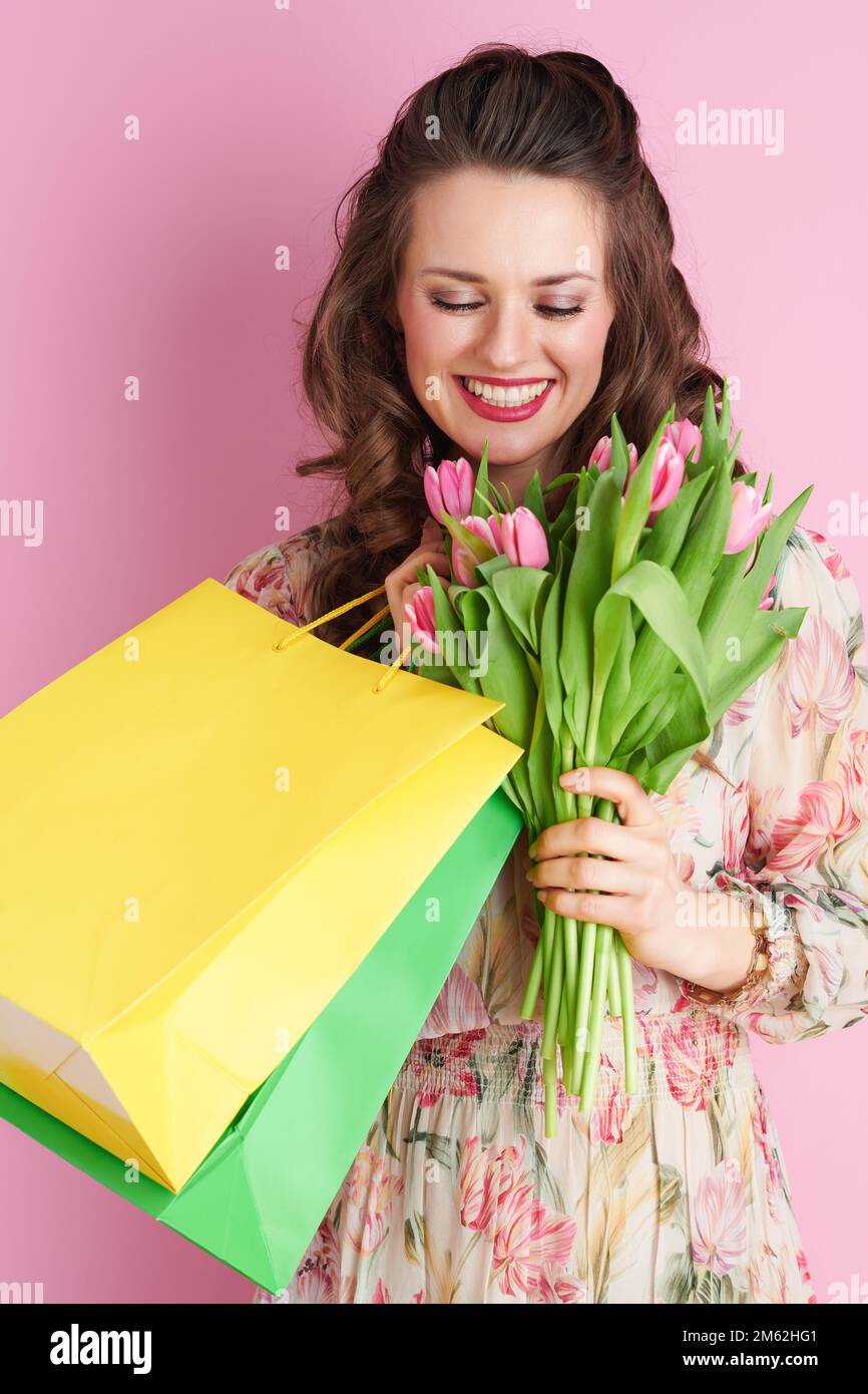 happy young woman with long wavy brunette hair with tulips bouquet and shopping bags isolated on pink background. Stock Photo