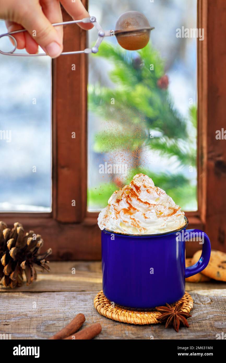 cinnamon powder from sieve flying on hot chocolate with whipped cream topping in ceramic cup on wooden table Stock Photo