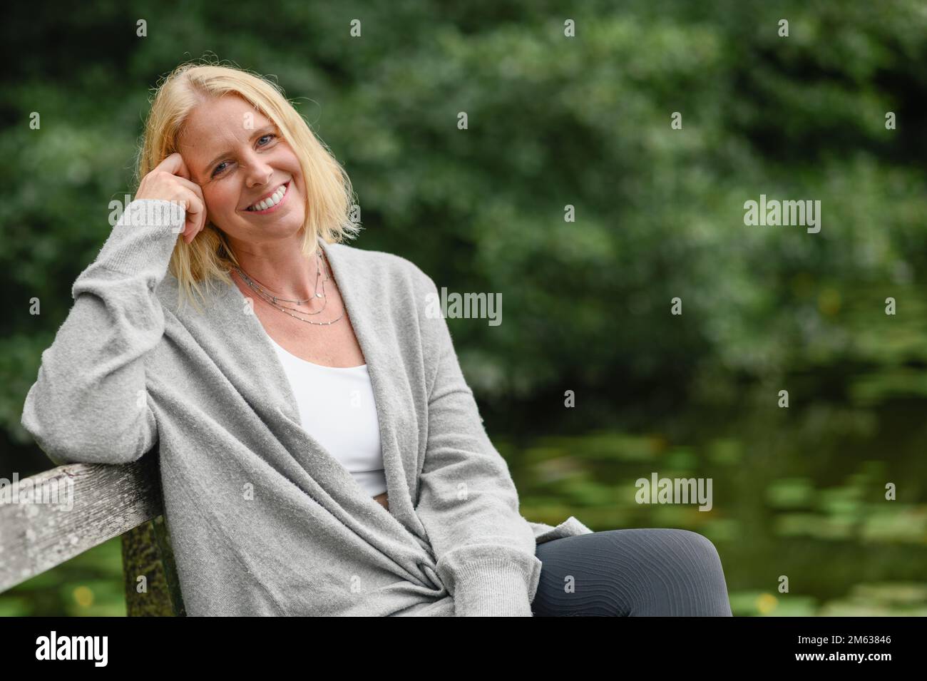 Positive middle aged blond haired female in gray cardigan leaning on hand while sitting on wooden bench near pond Stock Photo