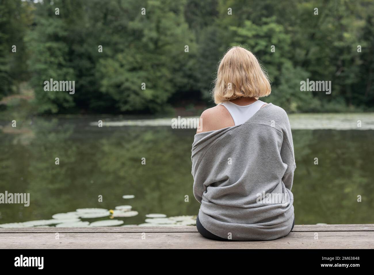 Back view of lonely blond haired female in casual clothes sitting on wooden pier near pond with green lotus leaves Stock Photo