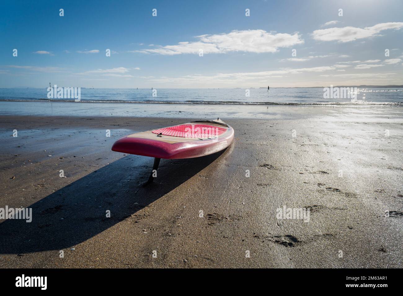 A stand up paddle board on a sandy beach with out-of-focus people paddle boarding in the sea. Auckland. Stock Photo