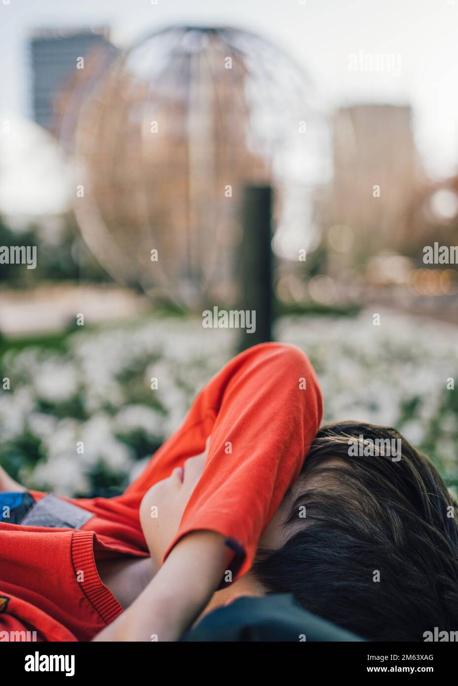 Side view of young boy lying in a city street park ground hiding face under arm covering eyes  Stock Photo