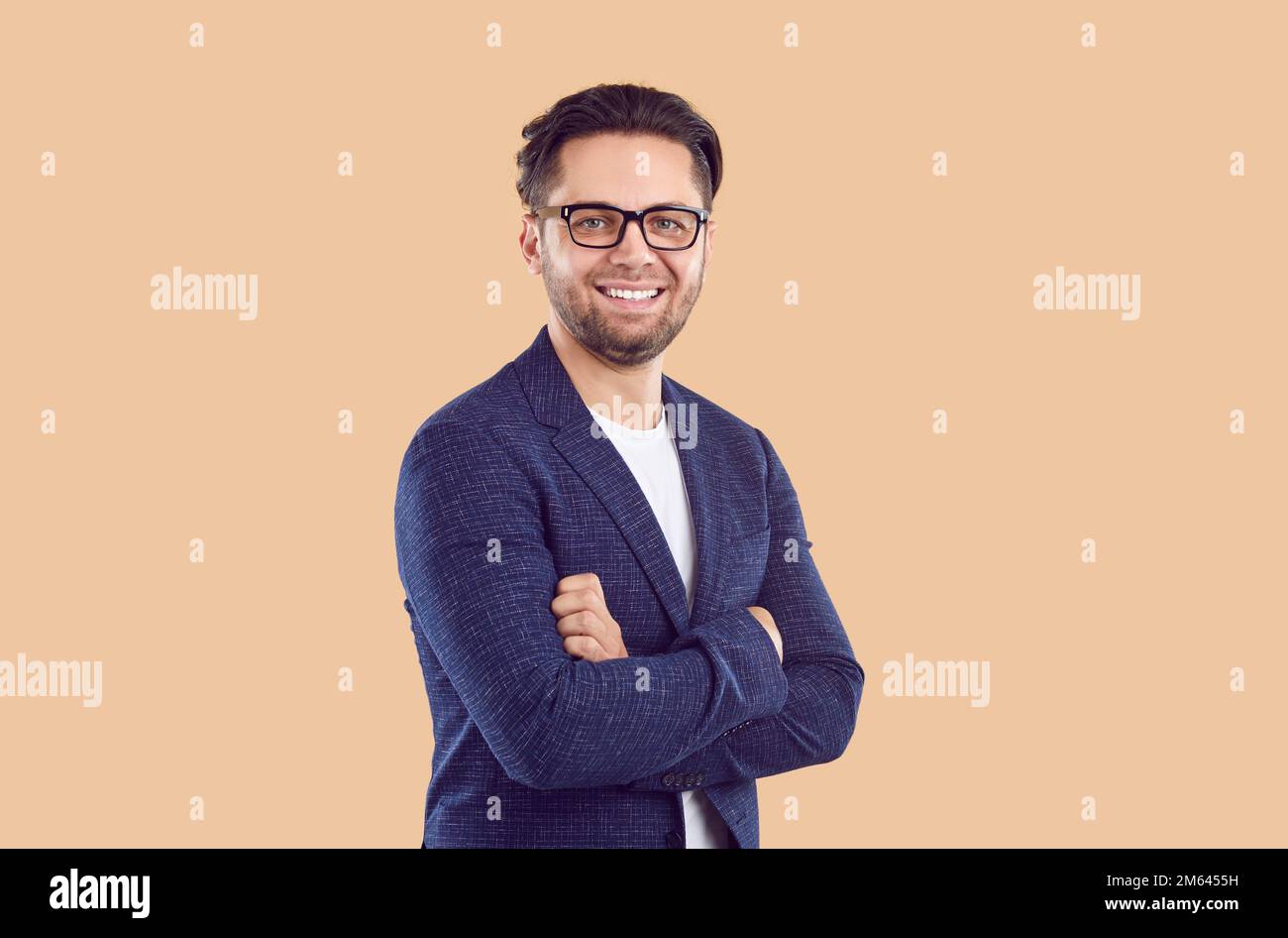 Smiling brunet man in blue jacket and glasses with crossing hands on chest on beige background. Stock Photo