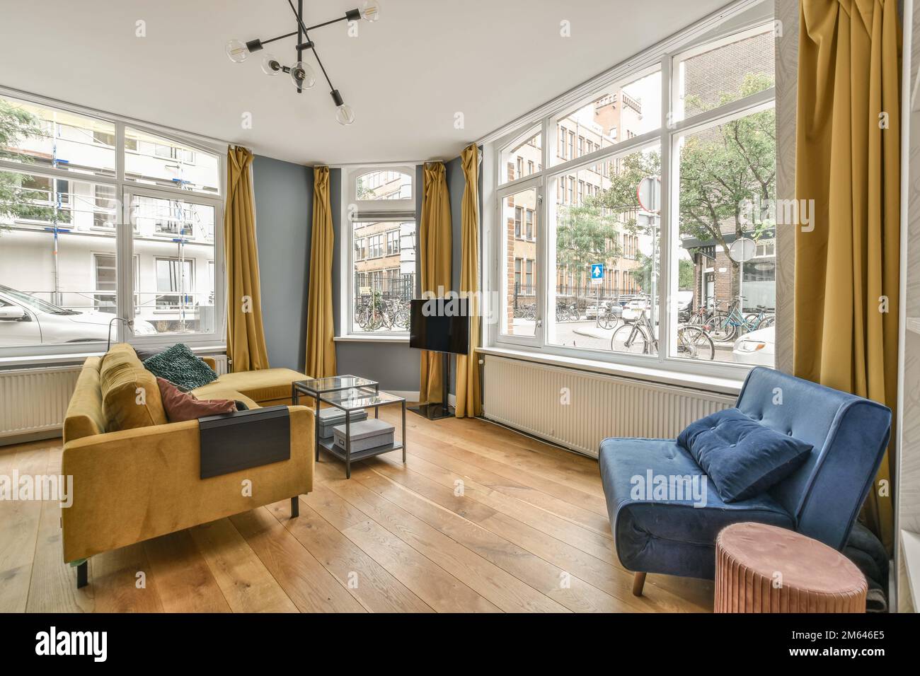 a living room with wood flooring and yellow drapes on the windows looking out onto an urban street view Stock Photo