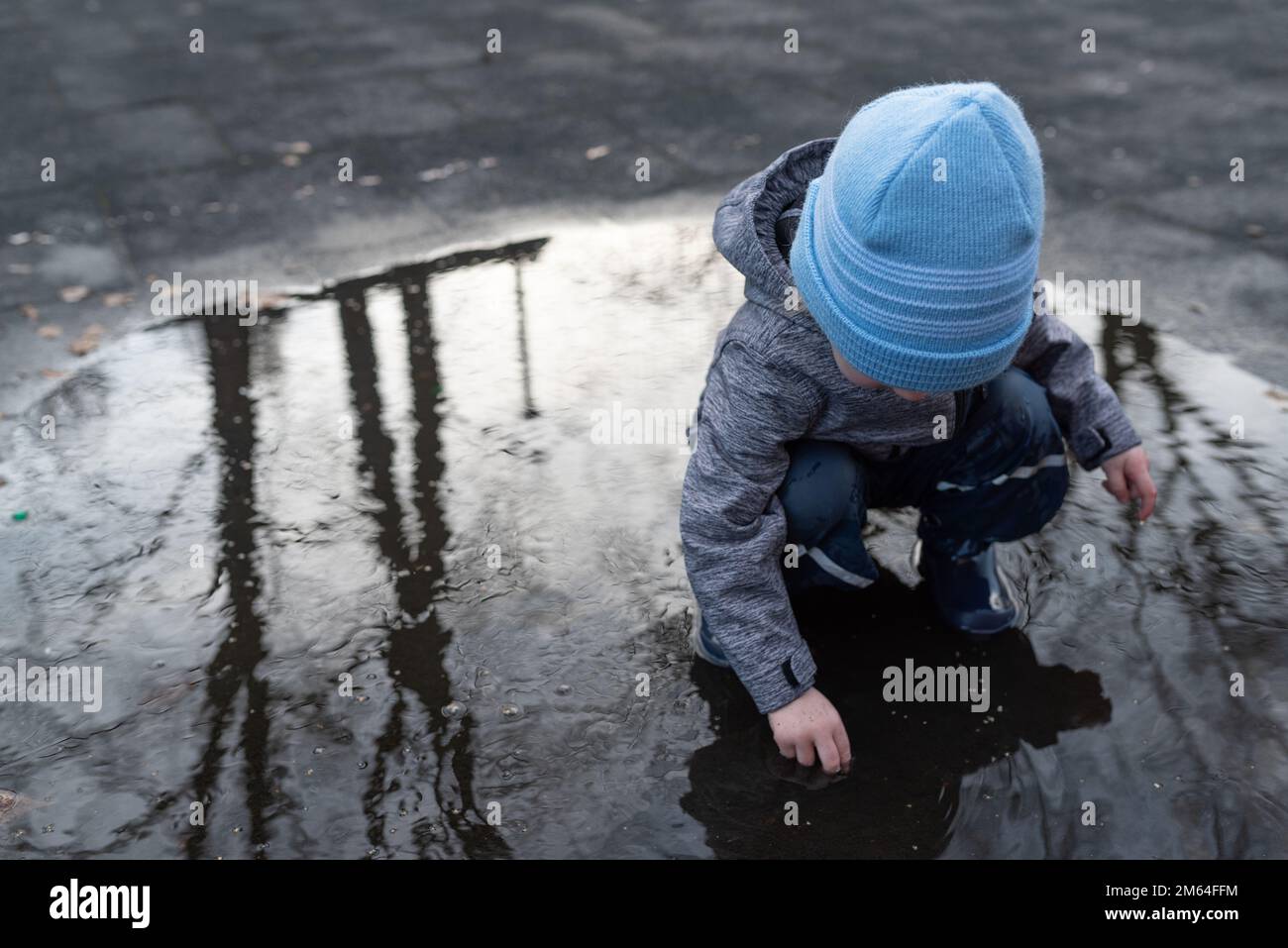 two year old boy in rain pants and rubber boots playing in puddle of water after rain shower Stock Photo