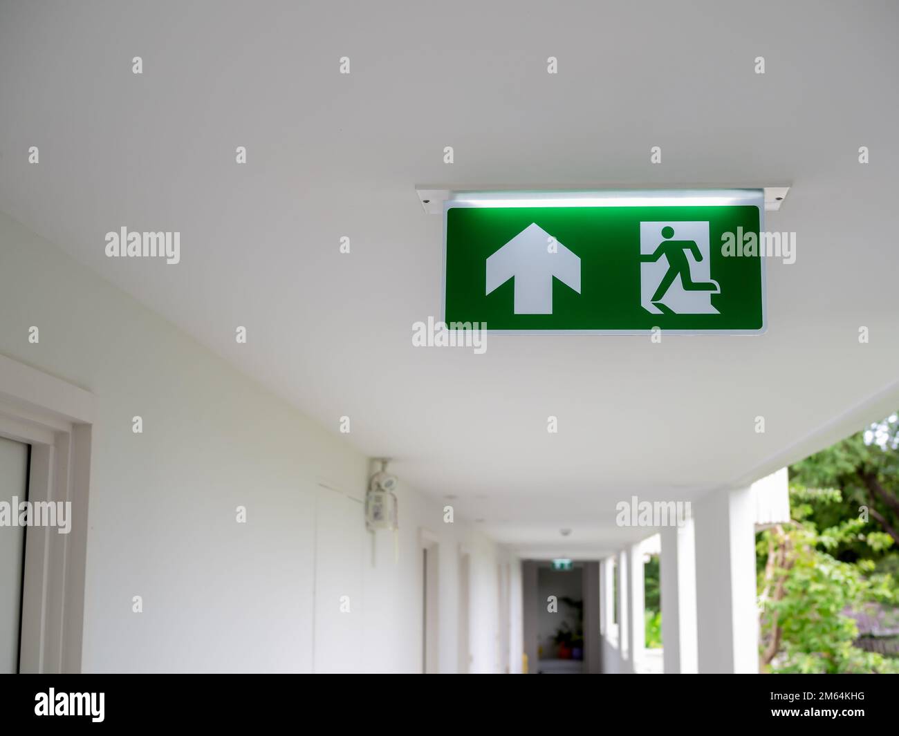 Green fire escape sign hang on the ceiling at the corridor in the white hotel building near the stairway. Emergency fire exit sign, warning plate with Stock Photo