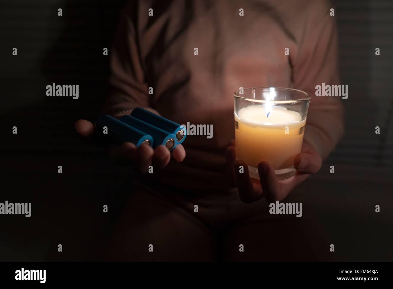 A girl in a dark room holds a candle and rechargeable batteries in her hands (close-up). Power outage concept. Blackout. Energy crisis Stock Photo