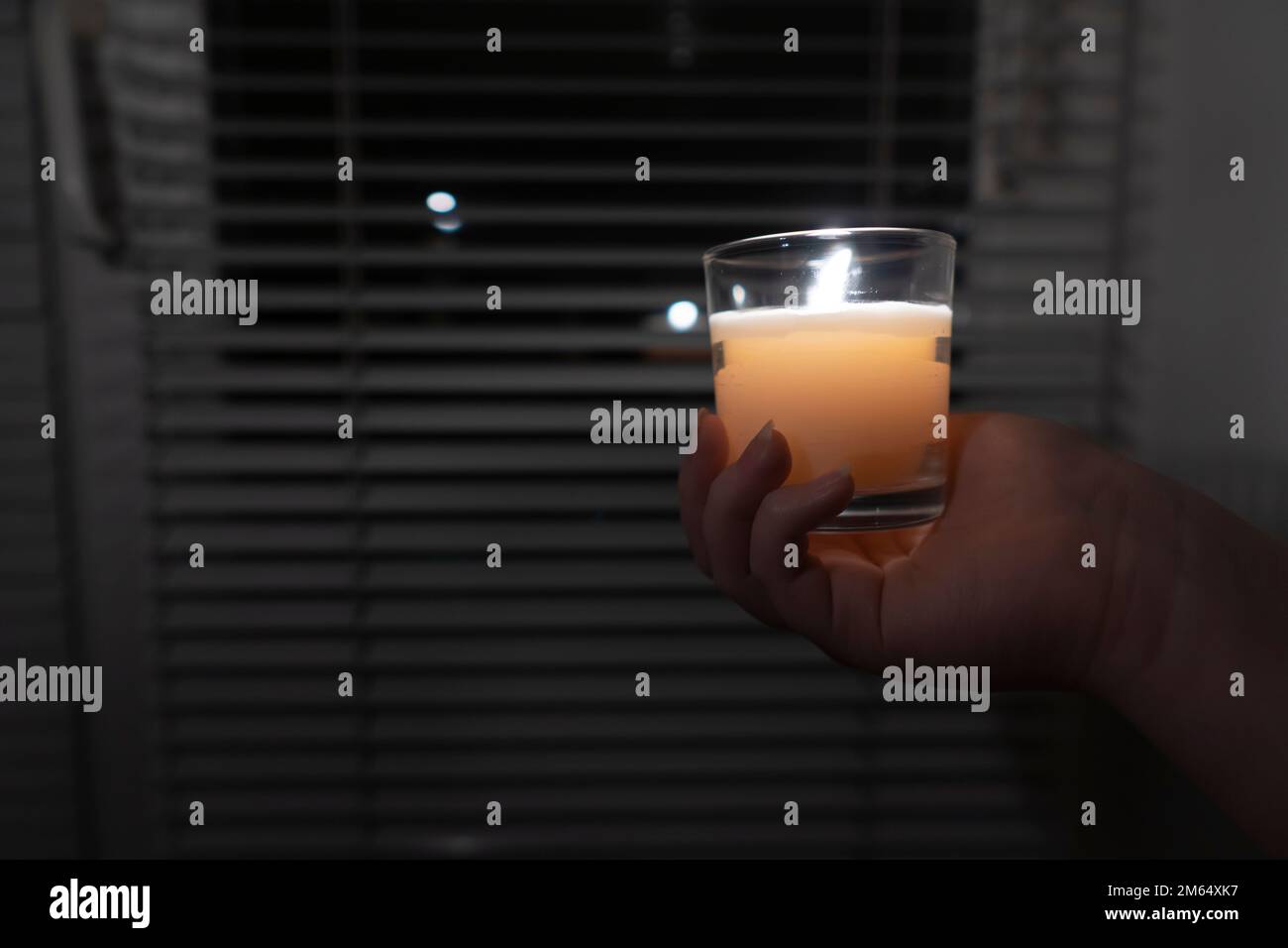 The girl holds a candle in a glass in her hand against the background of a window with blinds (close-up). Blackout. Energy crisis Stock Photo