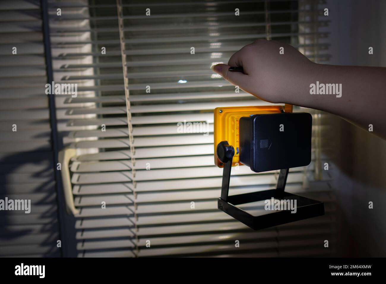 Power outage concept. Girl holds a bright flashlight in her hand against the background of a window with blinds (close-up). Blackout. Energy crisis Stock Photo