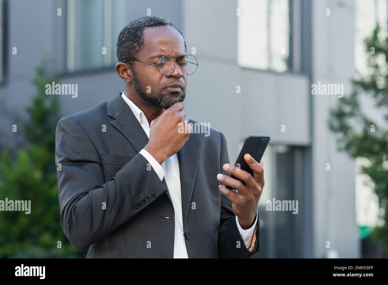 African american boss outside office building with phone in hands reading bad news online, man received notification about money loss and fraud, upset businessman. Stock Photo