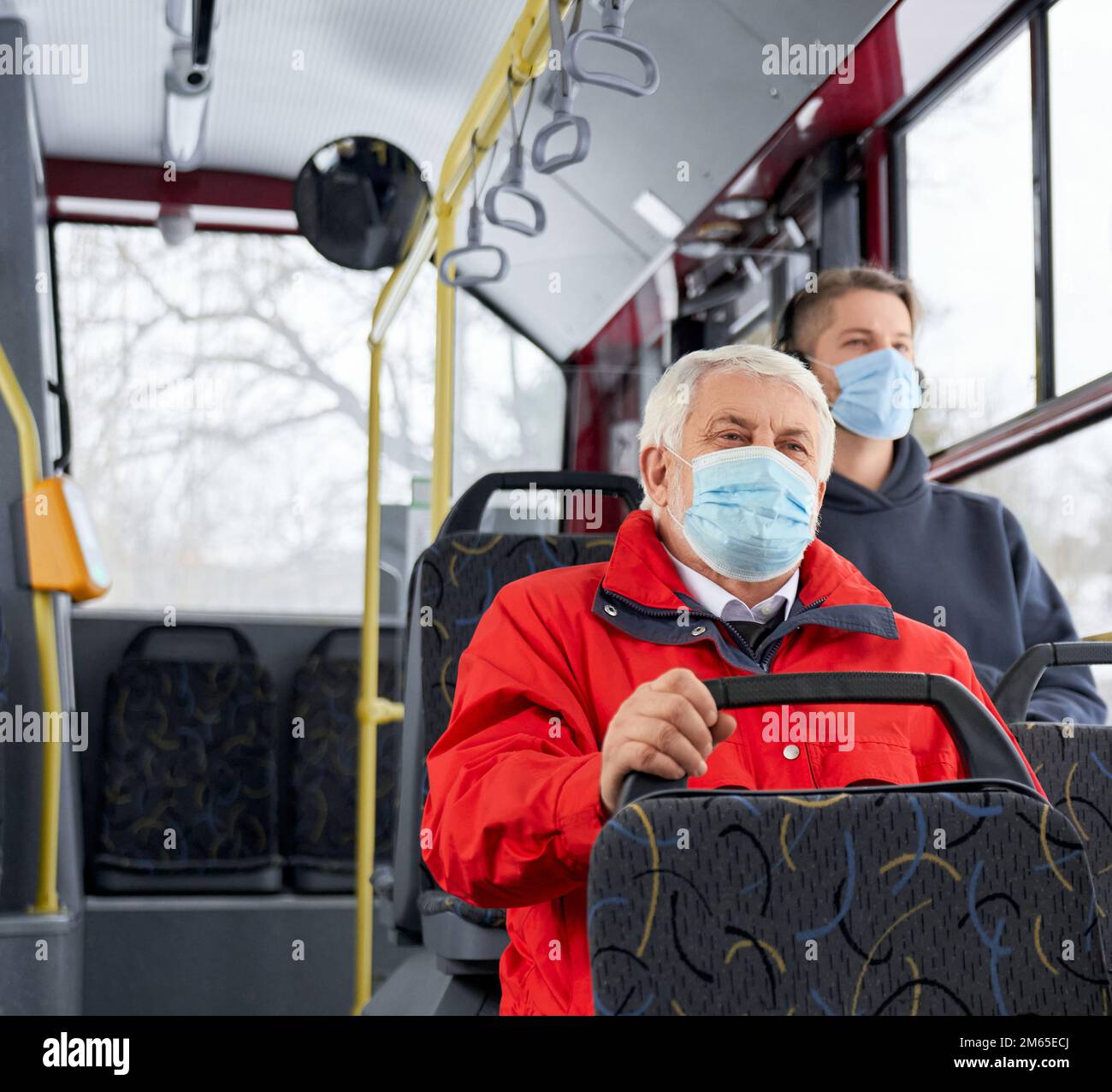 Passengers protecting by medical masks traveling by public transpost, sitting on bus. People going by bus home, old man and brunette man sitting, looking. Concept of pandemic. Stock Photo