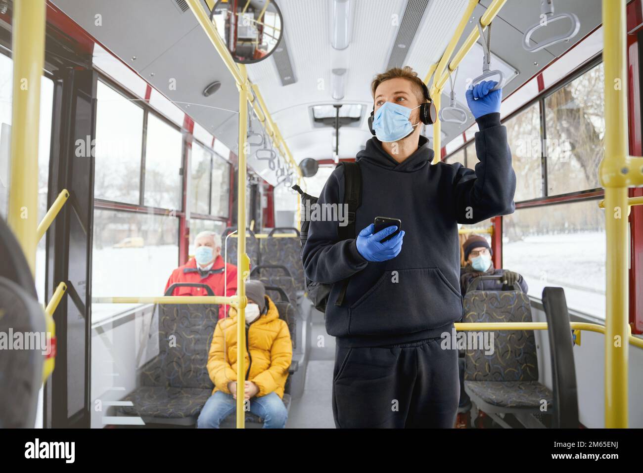 Front view of passengers going home by public transport, standing, sitting, wearing medical masks and gloves. Young man holding smartphone, paying. Co Stock Photo
