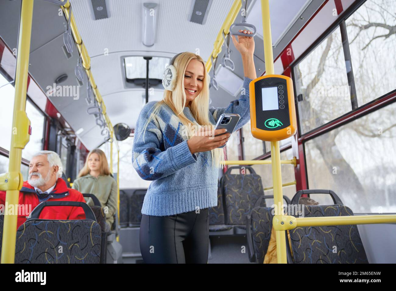 Portrait of fair long haired female wearing blue sweater and black trousers with earmuffs standing near bus payment terminal and performing contactles Stock Photo