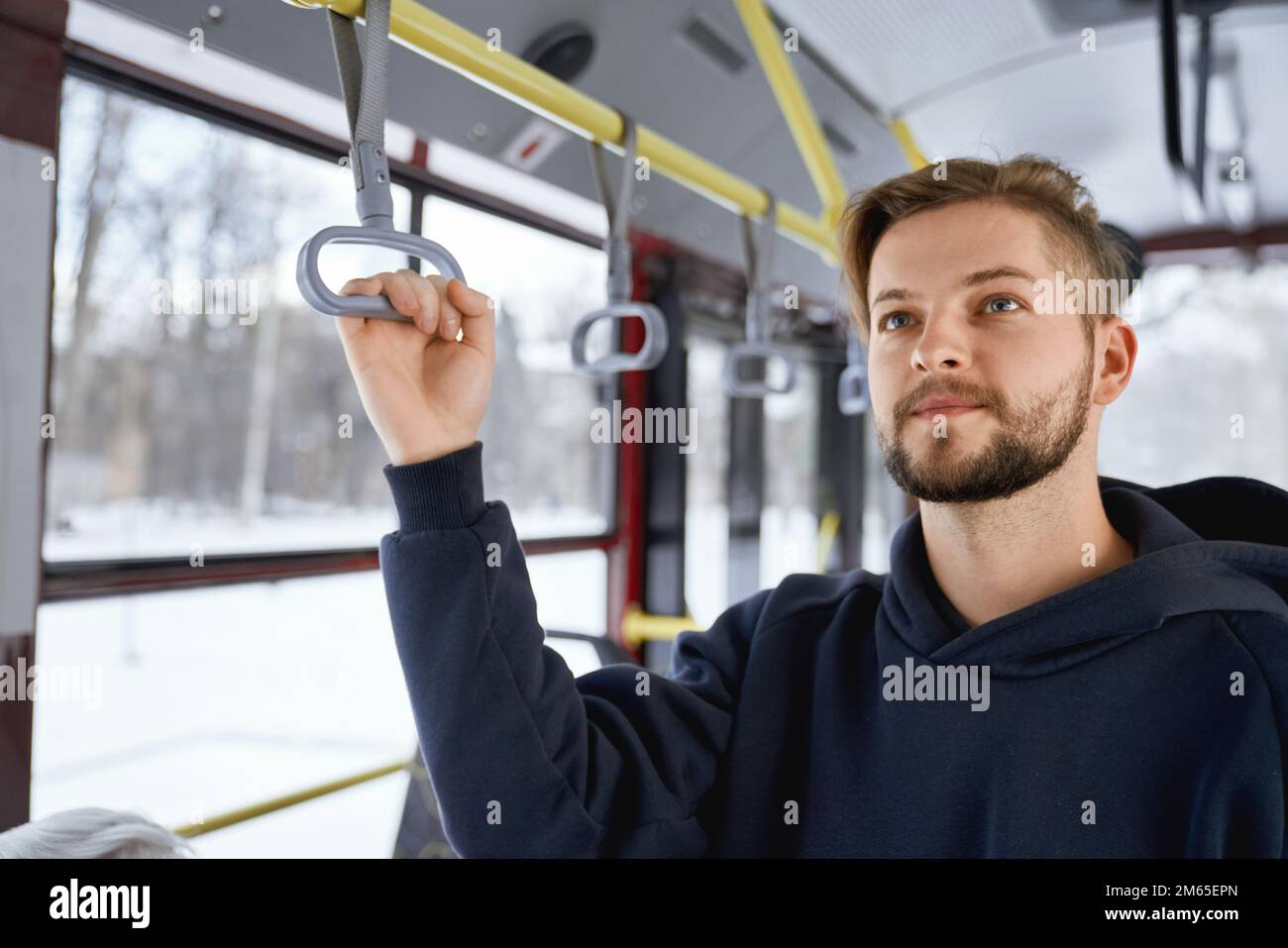 Front view of young male with beard going to work by public transport, standing on bus, waiting. Brunette man holding handle, wearing black khudi. Con Stock Photo