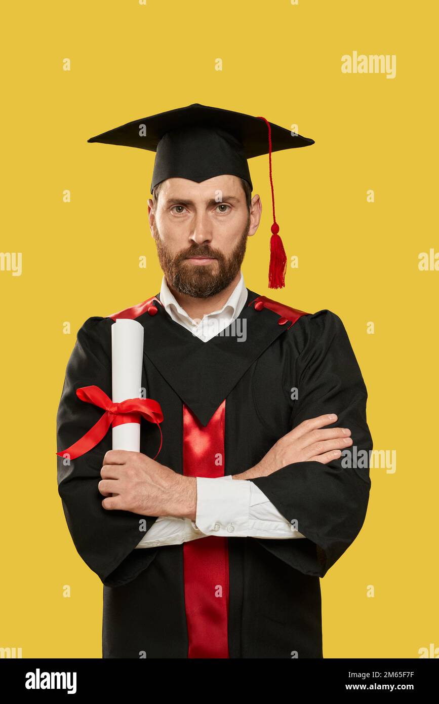 Front view of adult student standing with crossed hands, holding diploma. Handsome man with beard graduating, proud, successful, looking at camera Con Stock Photo