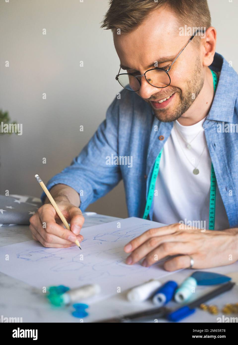Seamster with glasses is working at the table.DIY designer draws a sketch for a new costume project.Smiling man in a blue shirt makes a sketch Stock Photo
