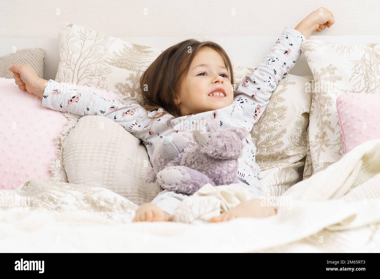 Happy little girl in sleepwear lying on bed and stretching with raised