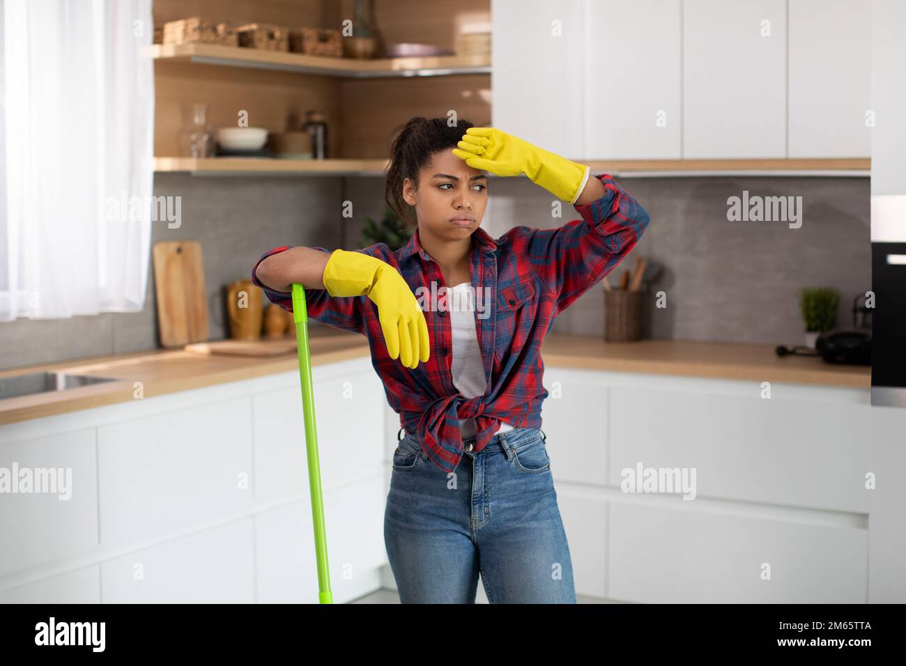Unhappy millennial african american housewife in rubber gloves wipes her forehead and mop floor Stock Photo