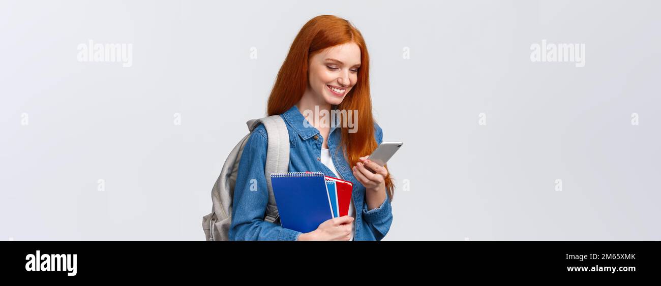 Lovely good-looking female with red hair, heading to college, holding backpack, notebooks smiling as answer friend message, chatting glancing mobile Stock Photo
