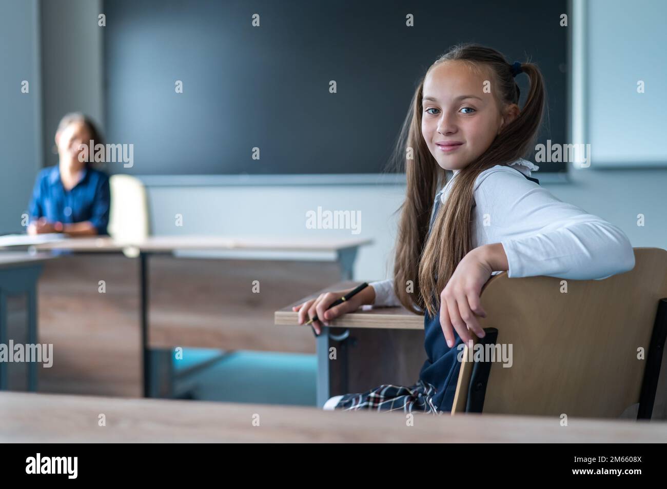 Caucasian girl and her teacher in the classroom. Schoolgirl turns around and looks at the camera.  Stock Photo