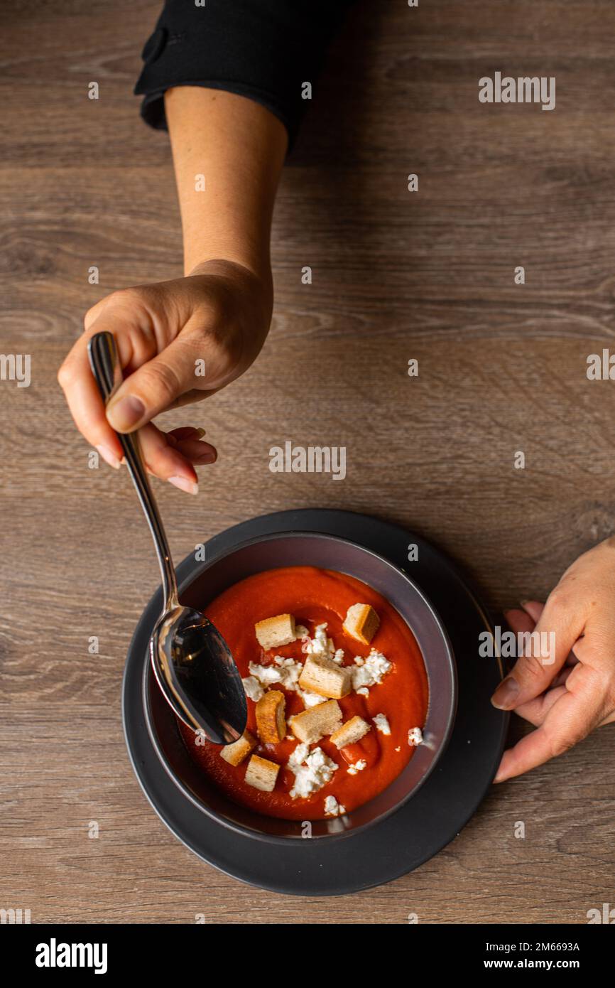 Vertical photo of cropped woman hands stir red tomato cream soup with soup crouton toasts on black plate on wooden table Stock Photo