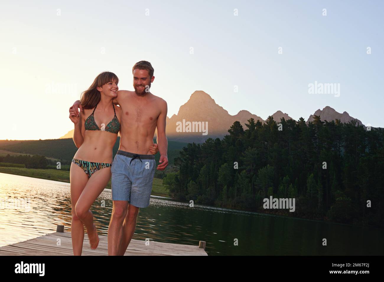 Love and laughter at the lake. an affectionate young couple in swimsuits walking on a dock at sunset. Stock Photo