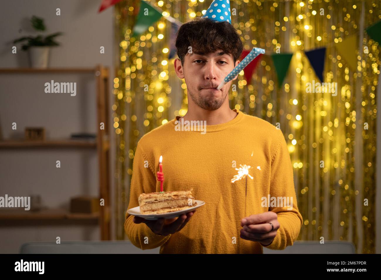 Cheerful young man in cone party hat with lit sparkler in hands celebrate his happy birthday to blow candle on his b-day cake during celebration at Stock Photo