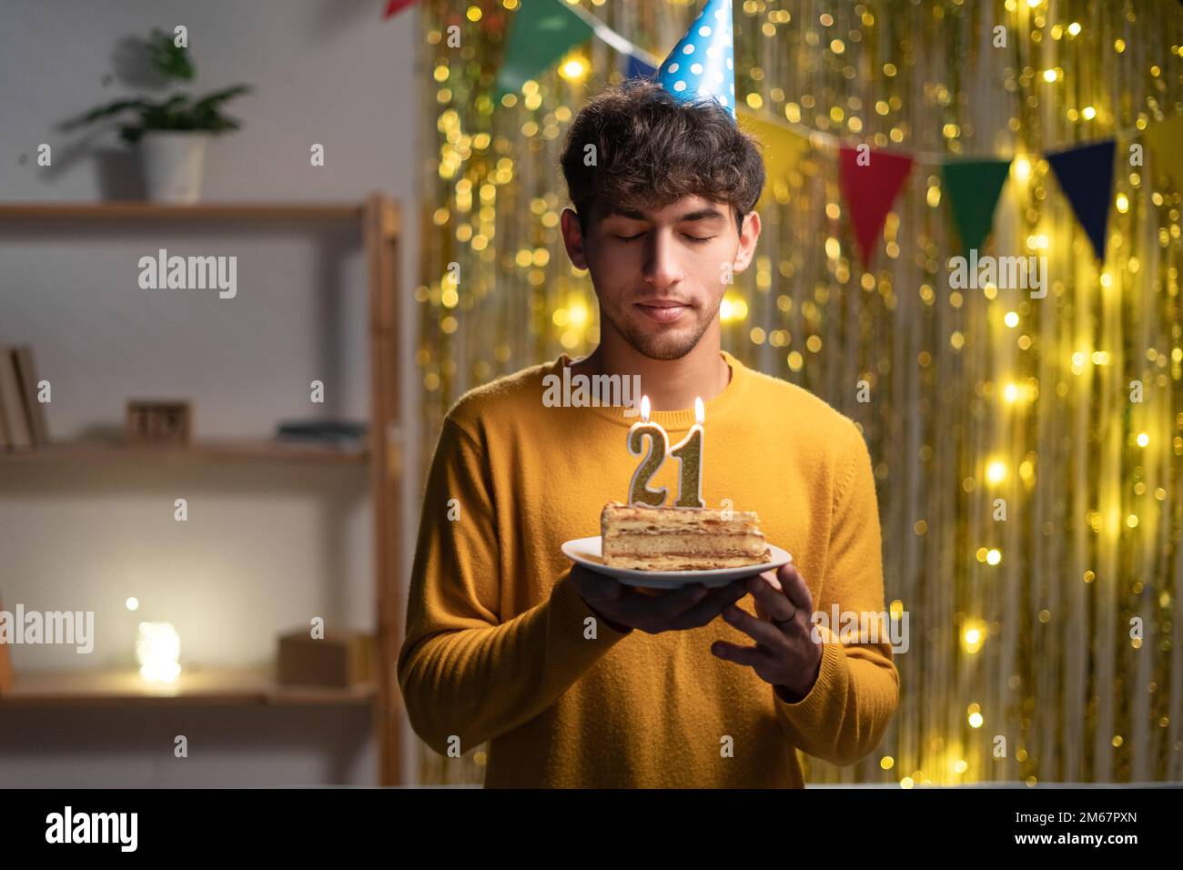 Guy celebrating 21st birthday at home, holding a cake with candles and making a wish Stock Photo