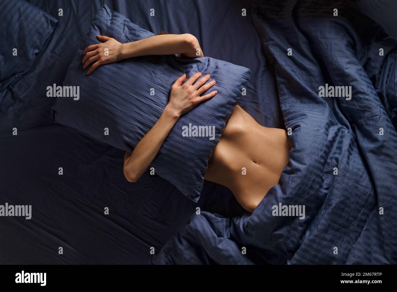 Concept of comfortable rest in bed, lazy weekend and introversion. Woman covers her face with pillow while lying in bed. Stock Photo