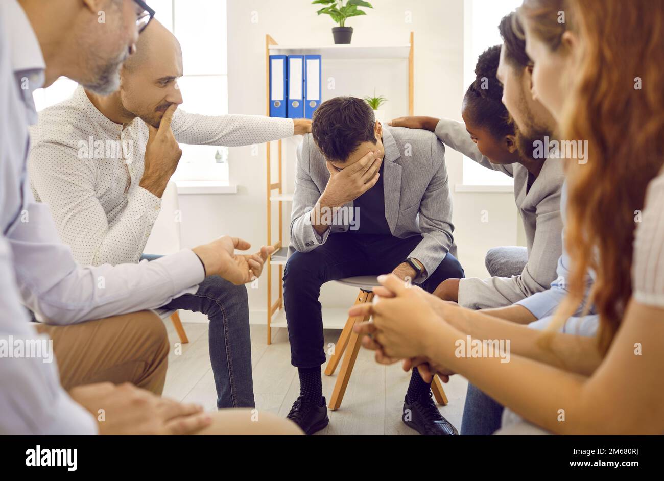 Sad man at support group therapy session sitting with head down and participants touch shoulders. Stock Photo