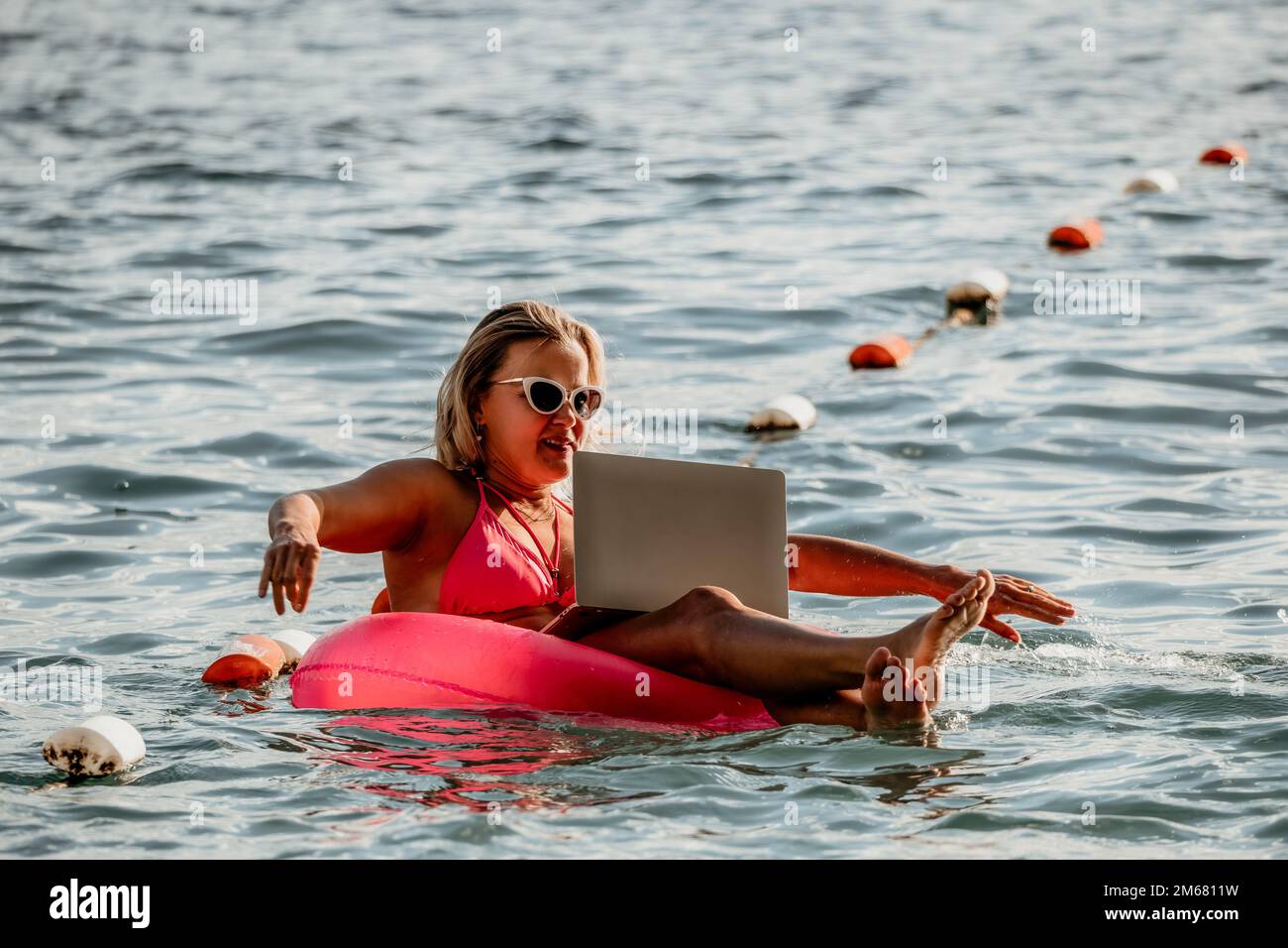 Woman works on laptop in sea. Freelancer, young blond woman in sunglases floating on an inflatable big pink donut with a laptop in the sea at sunset Stock Photo