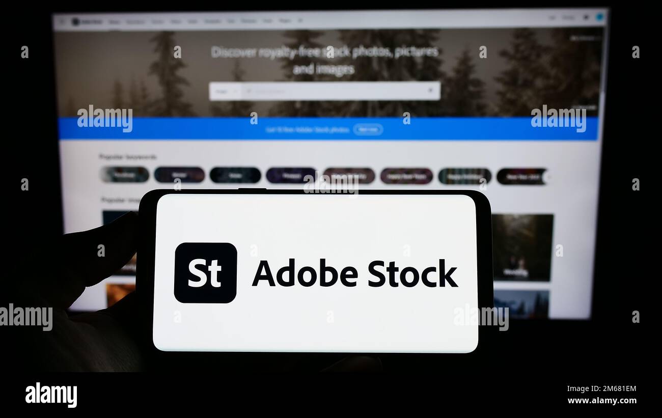 Person holding mobile phone with logo of American microstock agency Adobe Stock on screen in front of business web page. Focus on phone display. Stock Photo