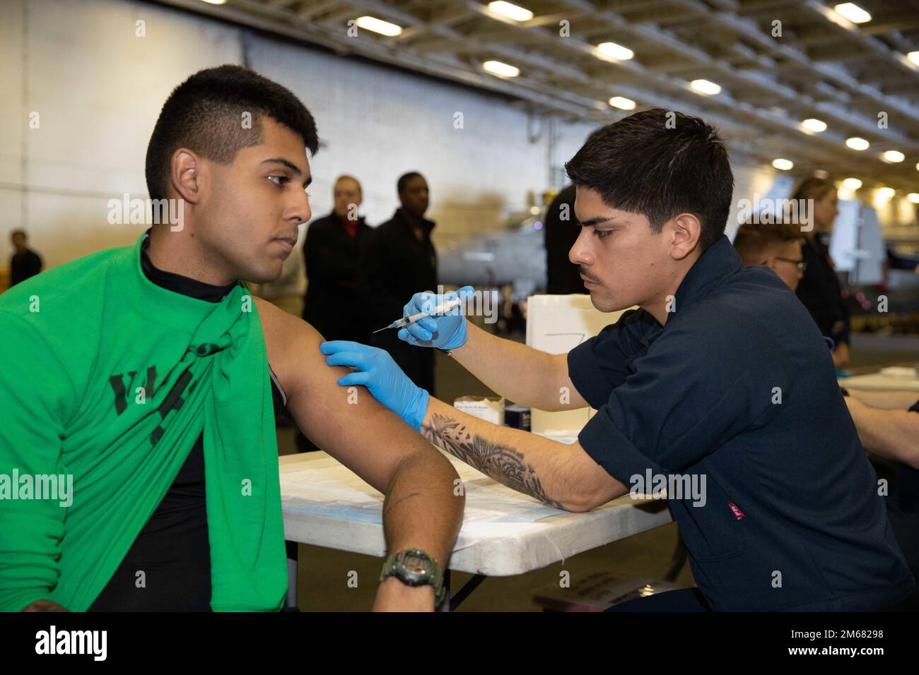 Hospitalman Michael Mino, right, from Newark, New Jersey, assigned to USS Gerald R. Ford’s (CVN 78) medical department, administers a vacine to Aviation Structural Mechanic Airman Apprentice Leonardo RamosRosado, from Sterling, Virginia, assigned to Electronic Attack Squadron (VAQ) 142, during a periodic health assessment in the hangar bay, April 15, 2022. Ford is underway in the Atlantic Ocean conducting carrier qualifications and strike group integration prior to operational deployment. Stock Photo