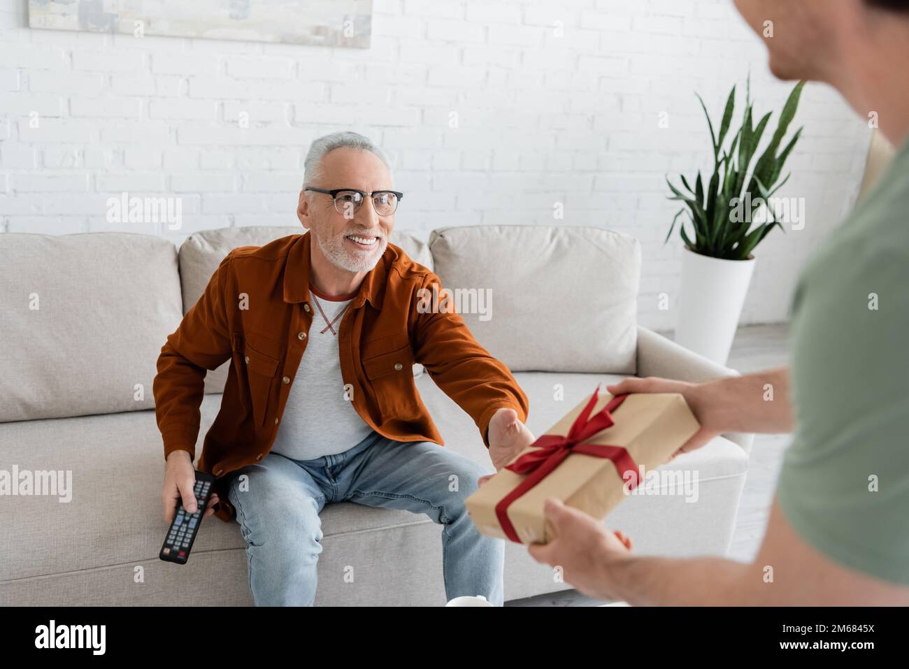 blurred man holding fathers day gift near smiling dad sitting on couch with tv remote controller,stock image Stock Photo