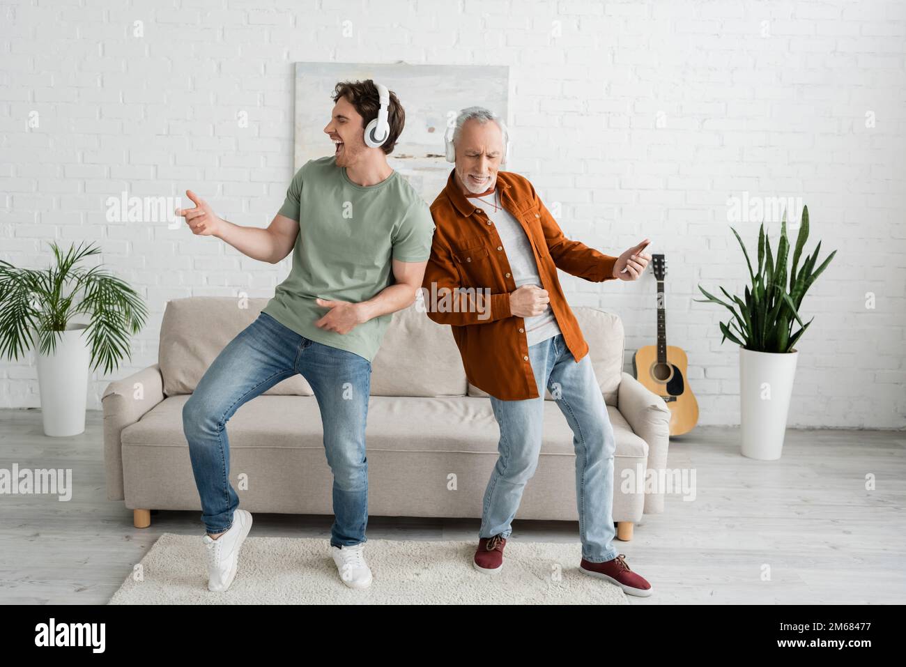 full length of young guy with senior dad in wireless headphones listening music and dancing in living room,stock image Stock Photo
