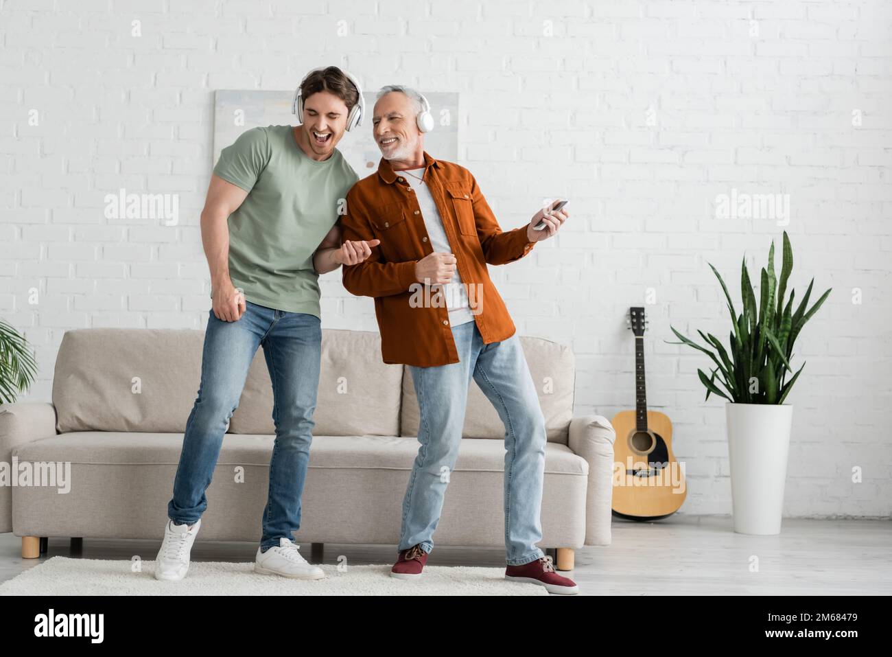 full length of joyful young man with mature dad in wireless headphones singing and dancing in living room,stock image Stock Photo