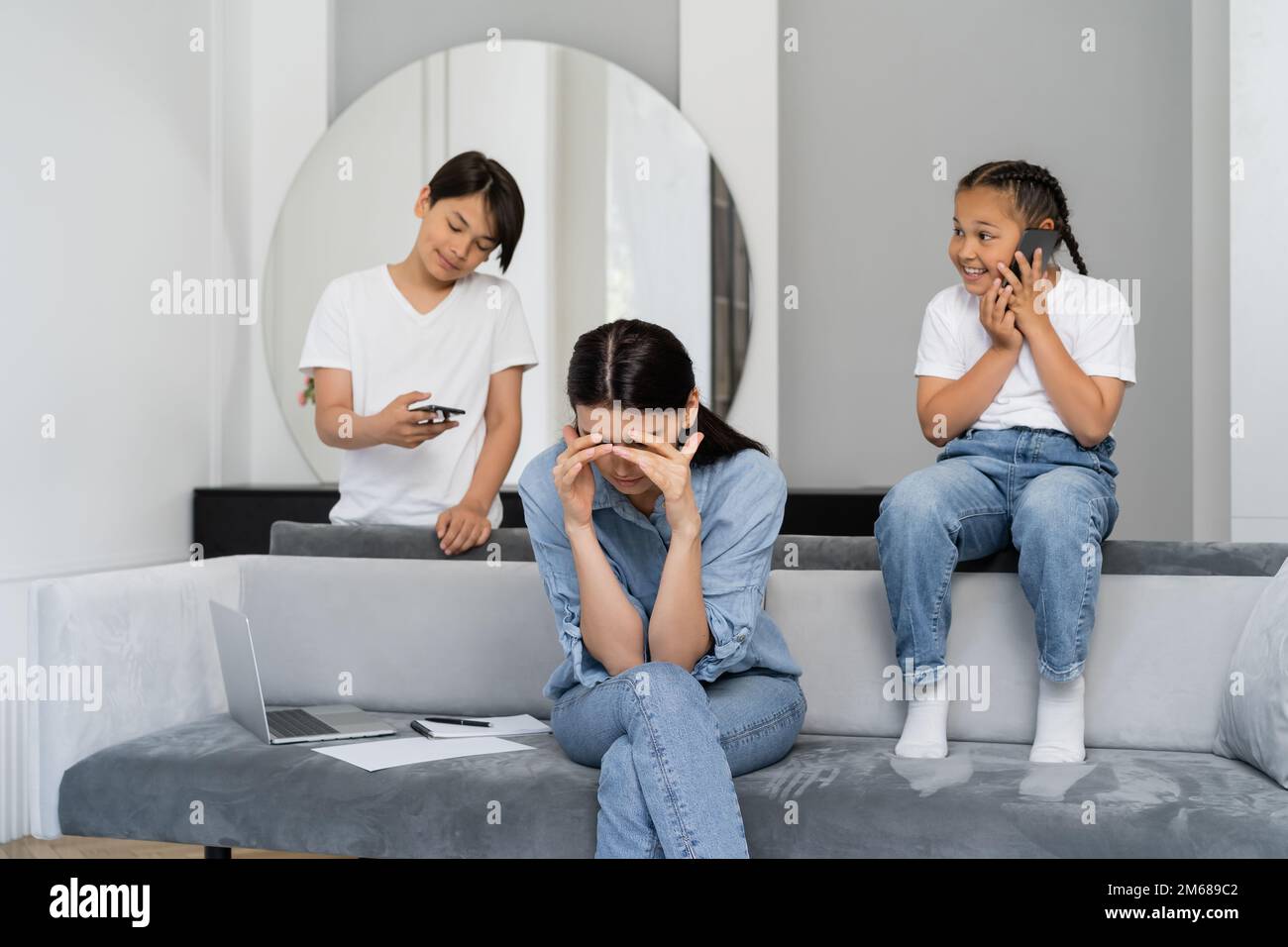 Tired mother sitting near laptop and asian kids using smartphones at home,stock image Stock Photo