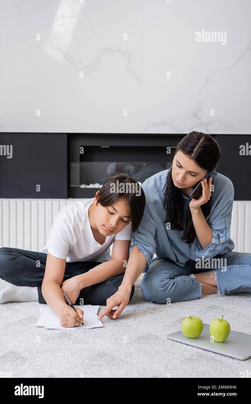 Woman talking on smartphone and pointing at paper while asian son doing homework at home,stock image Stock Photo