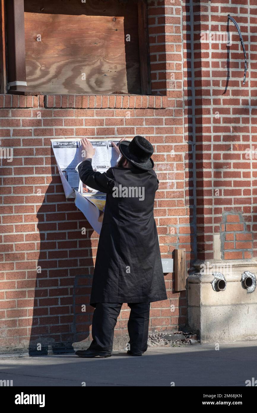 A Hasidic Jewish man hangs a sign with a schedule of religious events. Outside a synagogue on Lee Avenue in Williamsburg, Brooklyn, New York. Stock Photo