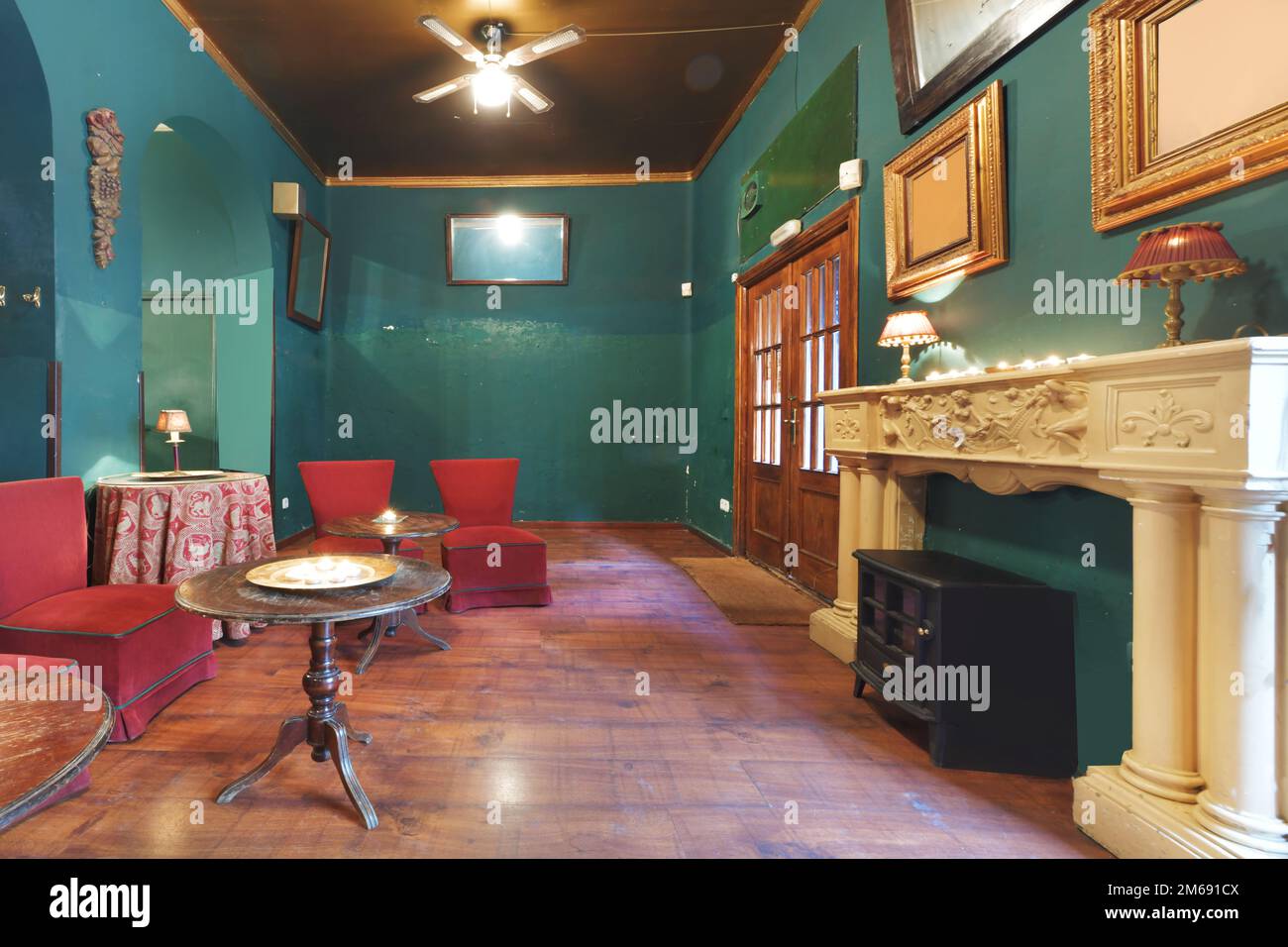 Hall of a vintage-style bar with green walls, a marble fireplace and armchairs upholstered in red fabric, round wooden tables and a fan with blades on Stock Photo