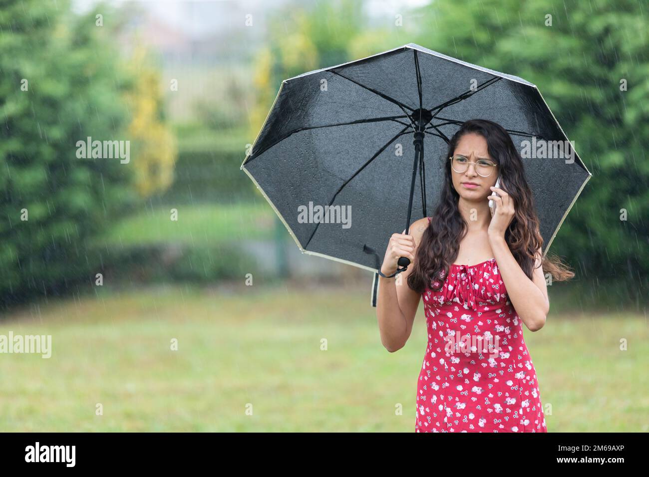 Ethnic woman with umbrella having phone argument in park on rainy day Stock Photo