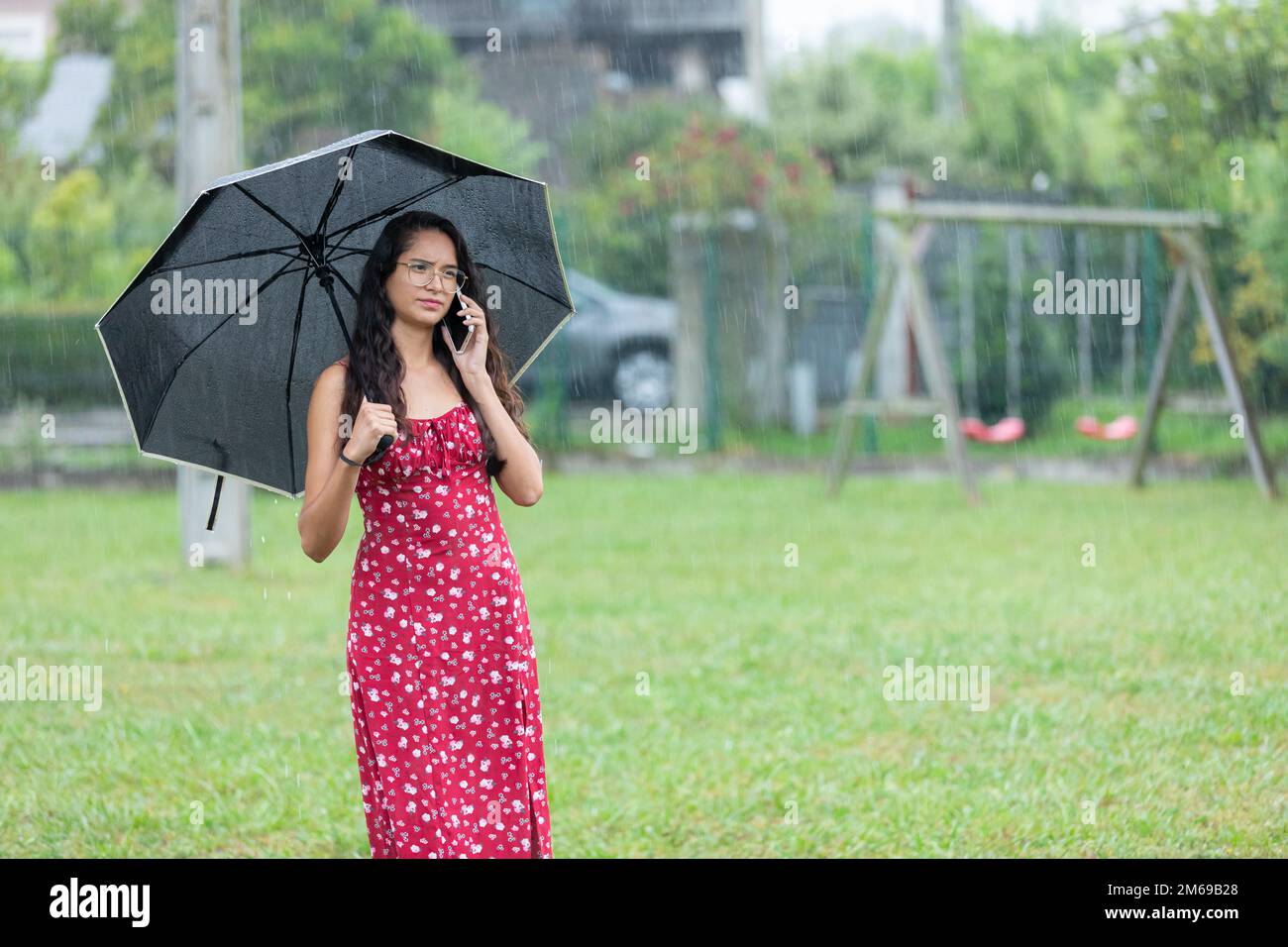Ethnic woman with umbrella having phone argument in park on rainy day Stock Photo