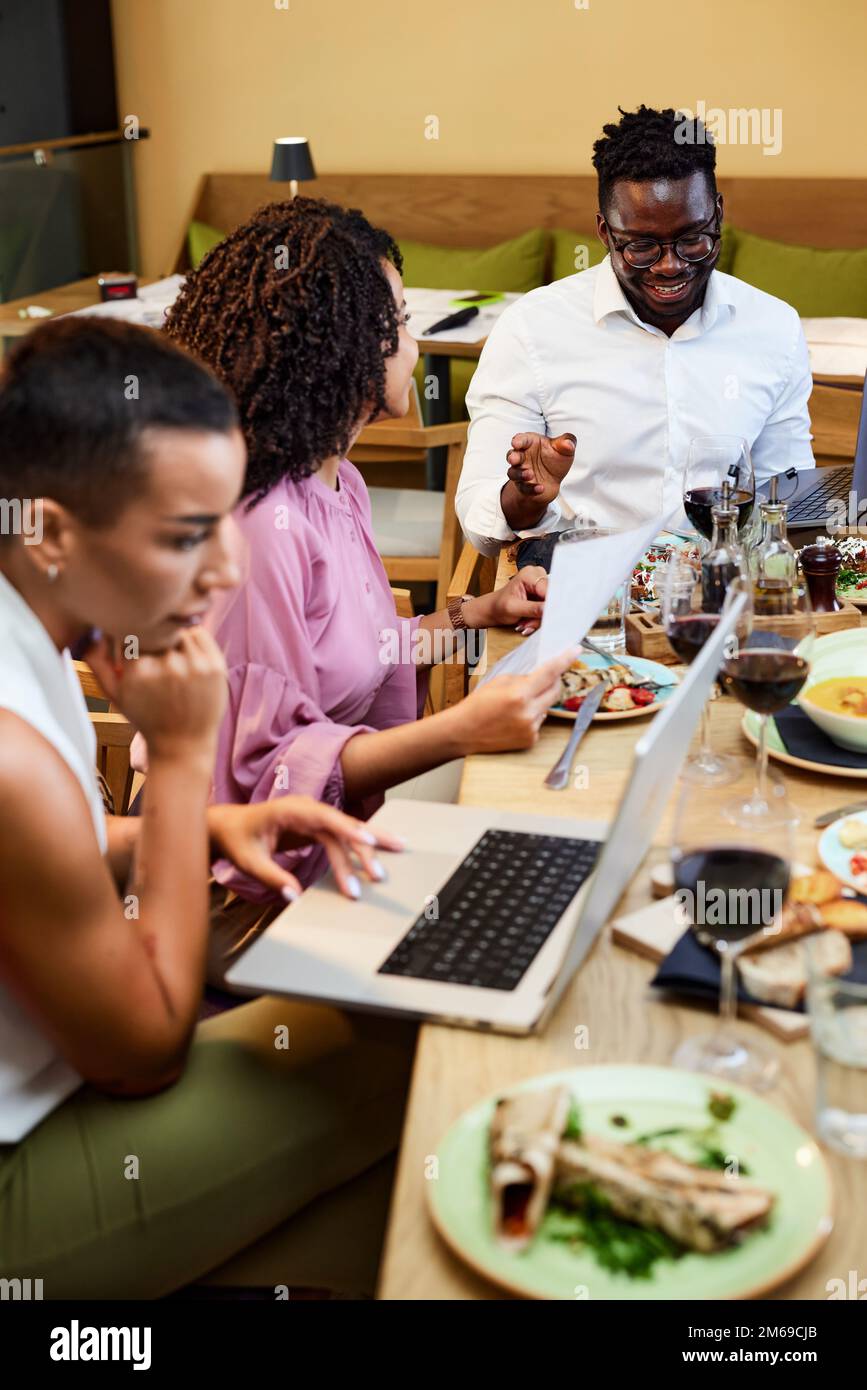 A cheerful group of multiracial colleagues having business lunch in a restaurant and working on the project. Stock Photo