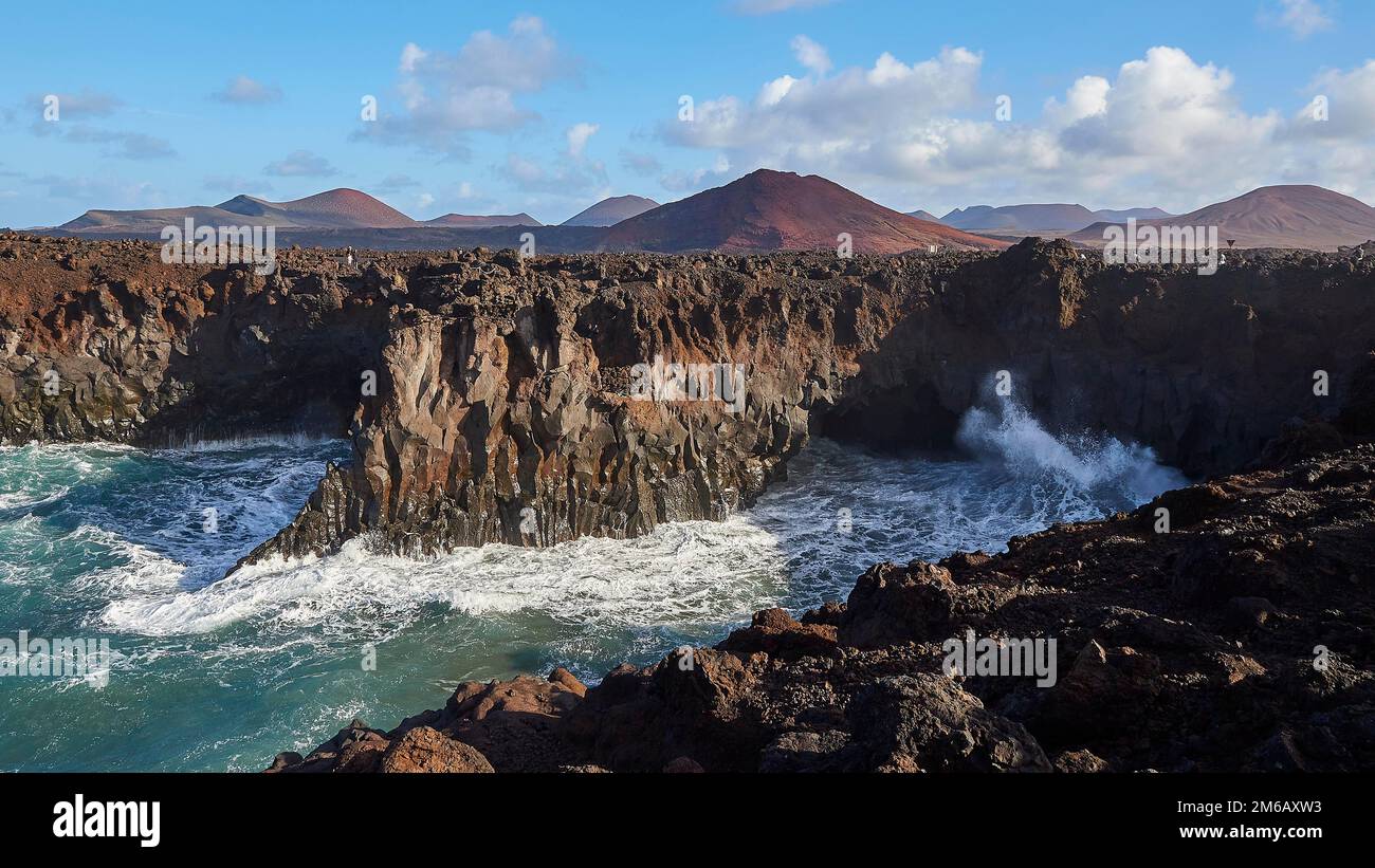 Los Hervideros, southwest coast, rugged volcanic coast, strong surf, sea caves, red lava hills, blue sky with white clouds, extremely clear air Stock Photo