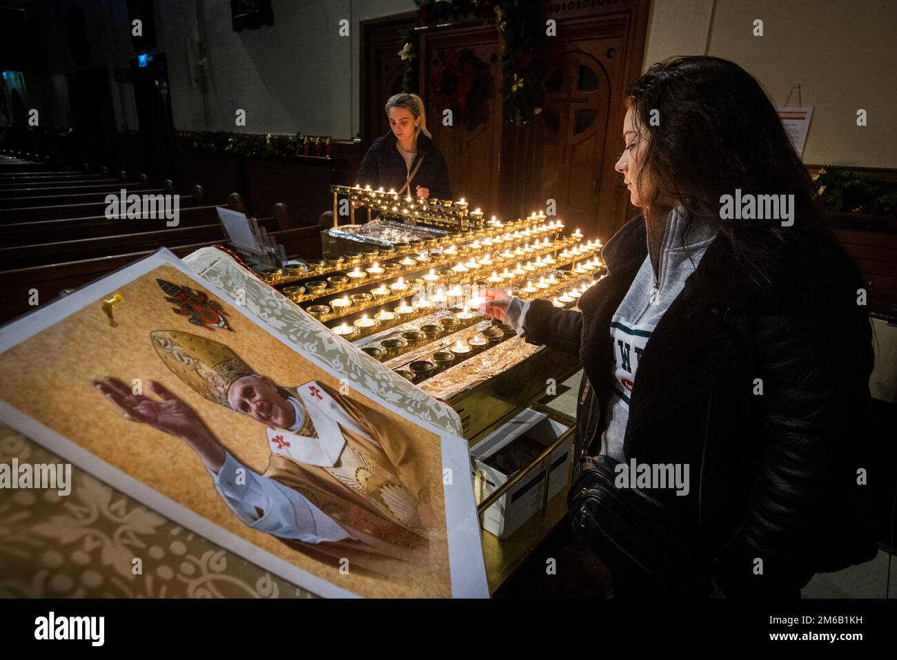 Lucy Knowles (right) with her cousin Amie Doak light candles beside an image of Pope Emeritus Benedict XVI at St Mary’s Church in Belfast, Northern Ireland. Benedict, 95, died on Saturday after 10 years in an extraordinary papal retirement lived out in a monastery in the Vatican Gardens. Picture date: Tuesday January 3, 2023. Stock Photo